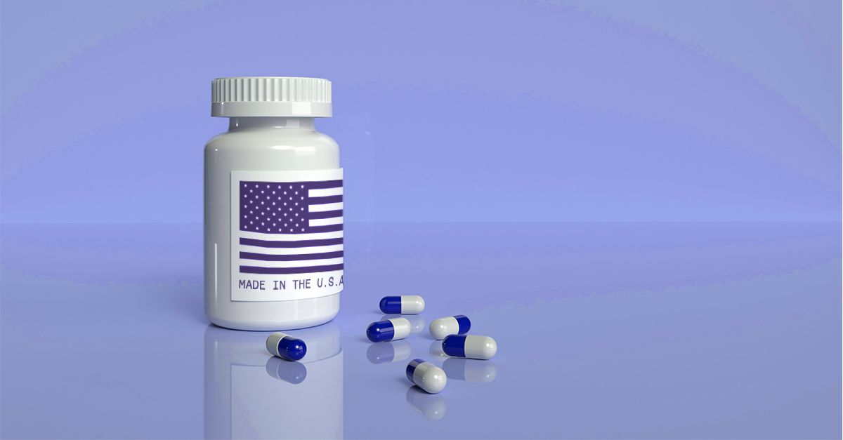 White prescription medicine bottle with an American flag label and the words “Made in the U.S.A.” printed below, symbolizing domestic pharmaceutical production. Several blue-and-white capsules are scattered on a glossy, reflective surface against a soft blue background, representing U.S. efforts to strengthen the pharmaceutical supply chain.