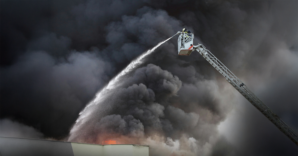 A firefighter in protective gear sprays water from an extended ladder platform toward thick, dark smoke and visible flames rising from a building fire.